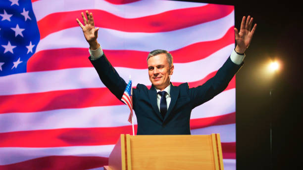 Friendly middle aged politician in suit gesticulating and talking with audience from podium against USA flag during election debate