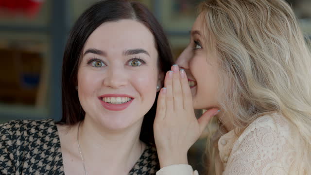Close-up face of excited young woman with surprised facial expression listening friend whispering on ear. Portrait of positive Caucasian women sharing secrets resting in cafe or restaurant.