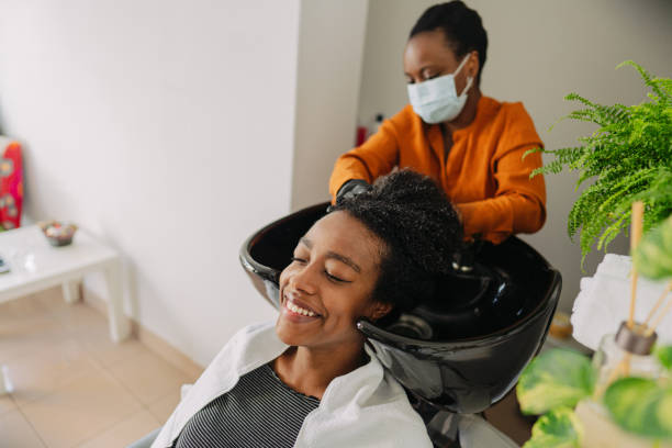 Visiting hair salon during pandemic Photo of a young hairdresser washing hair to a customer, while wearing protective equipment during the COVID-19 pandemic; visiting hair salons after a long period of lockdown. black woman washing hair stock pictures, royalty-free photos & images