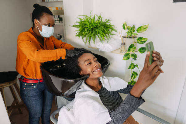 Visiting hair salon during pandemic Photo of a young hairdresser washing hair to a customer, while wearing protective equipment during the COVID-19 pandemic; visiting hair salons after a long period of lockdown. black woman washing hair stock pictures, royalty-free photos & images