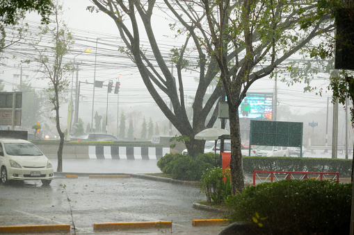 Heavy tropical rain storm in Bangkok and view from parking lot onto main road and junction at Nawamin Road. Cars are passing street.