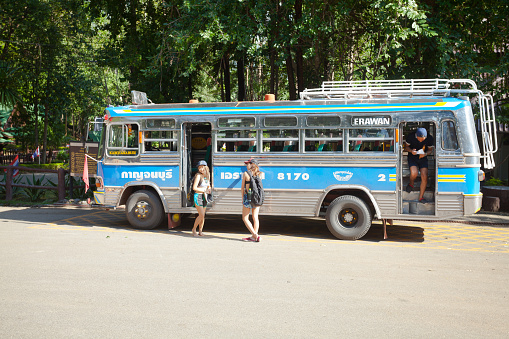 Young woman outside of public bus in Erawan national park in Kanchanaburi province. Scene is at parking lot and arrival point for bus transportation to Erawan waterfalls and park. A person is leaving the bus at back door