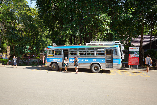 Young woman are posing for photos at public bus at Erawan waterfalls park. Bus is standing at arrival point in National park. Woman at left side is taking photos of women at bus. At right side a person is walking to buildings.