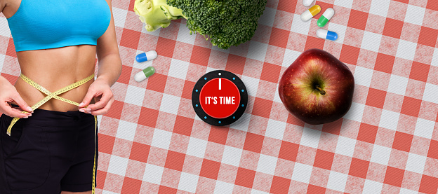 Healthy diet, exercise and weight loss concept for summer with a slender young woman measuring her waist against a checked red and white background with fresh broccoli and apple with disc with IT'S TIME text