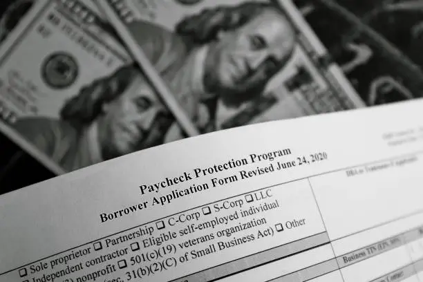 Photo of selective focus monochrome photo of paycheck protection program borrower application form revised, on a background of dollar bills and a chalk board with numbers