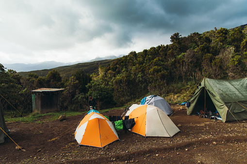 Scenic view of woman traveler walking near the dome tents on the camping ground in the beautiful green forest with view of the mountain peaks under dramatic sky on Mount Kilimanjaro, Tanzania