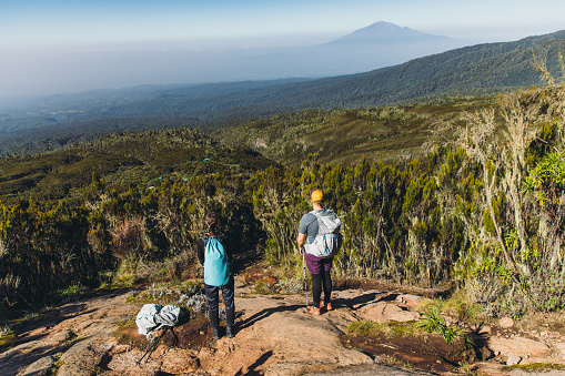 Woman and man with backpacks relaxing after the hiking trip on Mount Kilimanjaro looking at the beautiful blue mountain and the green valley
