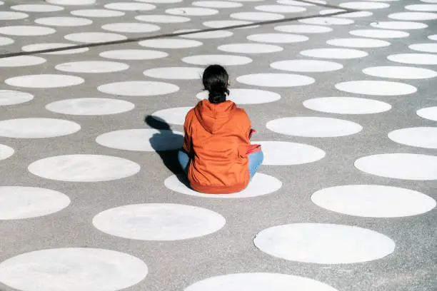 rear view on girl in orange hoody sitting cross-legged on white spotted town square in Berlin under afternoon sun