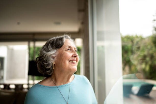 donna anziana che guarda attraverso la finestra di casa - senior adult 70s female women foto e immagini stock