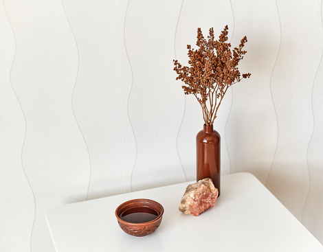 Interior composition. Clay cup of tea, natural stone, a clay vase with brown dry flowers on a white coffee table against a white wall.