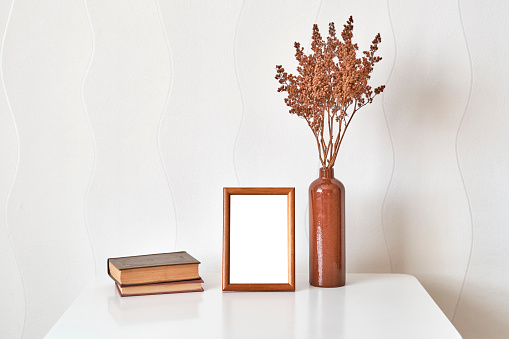 Interior composition. Wooden brown frame mock up, clay vase with brown dried flowers, stack of two old small books on a white coffee table against a white wall background.