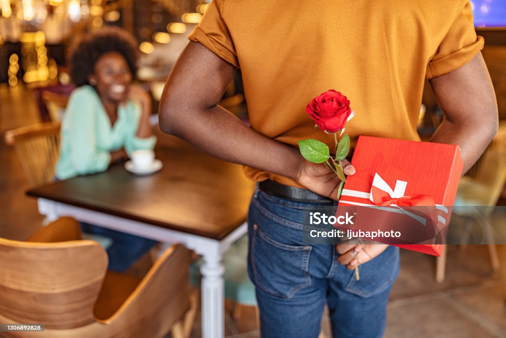 Valentines day Man hiding red rose behind his back and gift for his smiling girlfriend while sitting in restaurant. Valentine's Day - Holiday Stock Photo