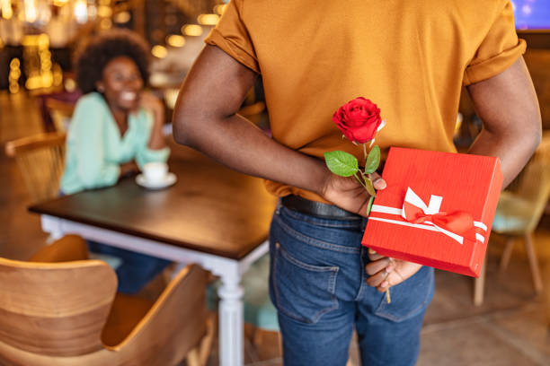 Valentines day Man hiding red rose behind his back and gift for his smiling girlfriend while sitting in restaurant. anniversary gifts stock pictures, royalty-free photos & images