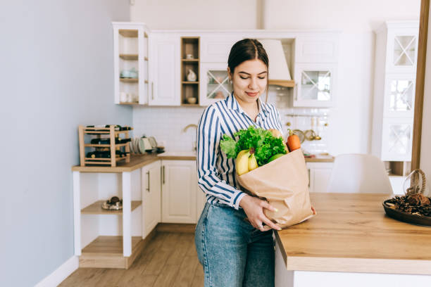 young smiling caucasian woman hold eco shopping bag with fresh vegetables and baguette in modern kitchen. - papel de pão imagens e fotografias de stock