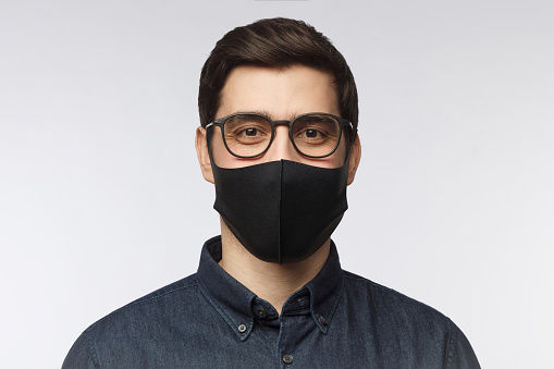 Young man wearing denim shirt, glasses and black mask standing isolated on gray background
