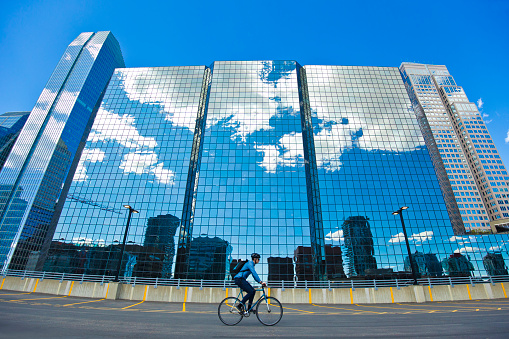 A male bicycle commuter rides his bike through a parking lot on his way to work in a big city. He rides a single-speed or fixed-gear bicycle with just a front brake. He wears a cycling helmet and carries a bicycle messenger-style backpack. He rides past the office buildings that reflect the clouds in the sky.