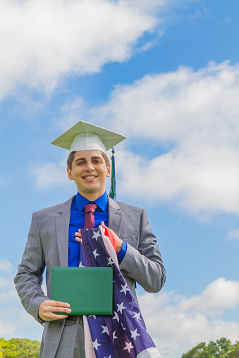 Portrait of Cheerful Proudly happiness handsome latin male student wearing a grey suit, blue shirt, red tie, gown and a hat, attending his Graduation Ceremony, holding a Graduating Certificate from High School College / University with hand on his heart with the US USA American Flag, standing and posing at the garden in front of school building in Miami, Florida, United States of America, in a blue summer midday, looking directly to camera on his graduation day.\n\nEducation, graduation and people concept. \nReal People doing real things.