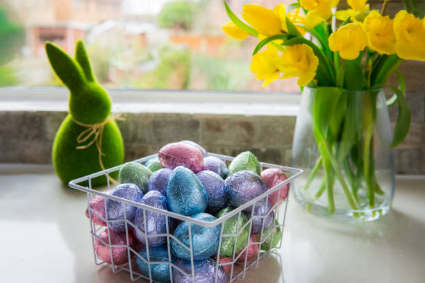 cesta de metal con huevos de pascua de chocolate en papel de aluminio de color, ramo de tulipanes amarillos y flor de narcisos y conejo verde pascua en la mesa de la cocina blanca cerca de la ventana. composición festiva de primavera. - daffodil easter egg hunt easter easter egg fotografías e imágenes de stock