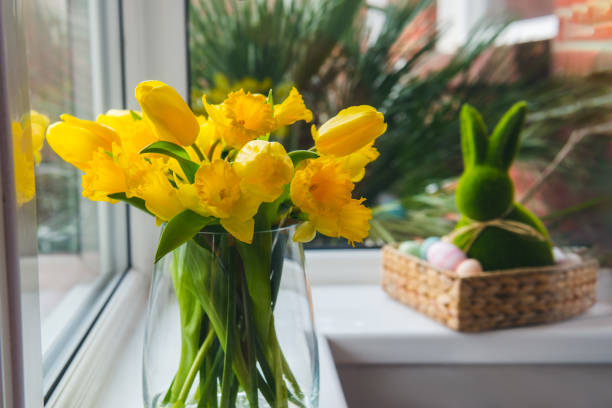 istock Bouquet of fresh spring tulips and daffodils flowers on the windowsill with Easter bunny rabbit in straw basket with colored eggs on background. Happy Easter festive card. Selective focus. Copy space. 1306912059