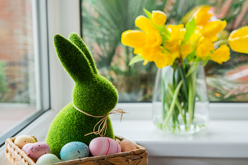 Easter bunny rabbit statuette in straw basket with colored eggs on the windowsill with fresh spring tulips and daffodils flowers bouquet on the background. Happy Easter. Selective focus. Copy space