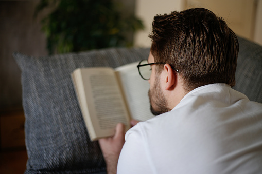 Young businessman in casual clothes and eyeglasses reading a book while lying on couch at home
