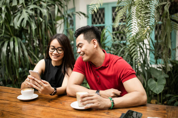 beautiful adult couple at restaurant shopping online while man holds credit card both smiling - using phone garden bench imagens e fotografias de stock