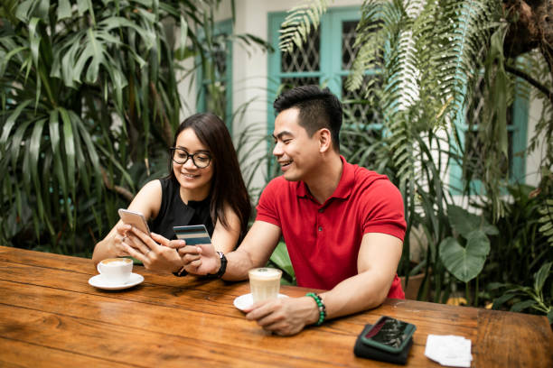 beautiful adult couple at restaurant shopping online while man holds credit card both smiling - using phone garden bench imagens e fotografias de stock