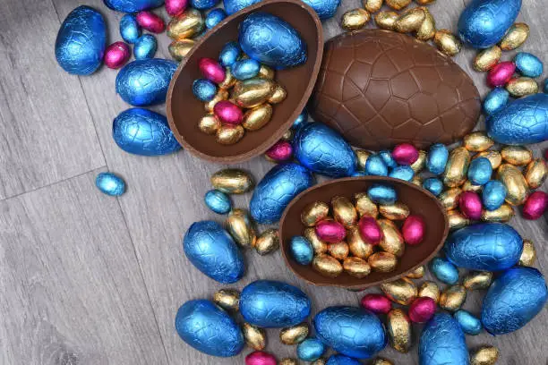 Pile or group of multi colored and different sizes of colourful foil wrapped chocolate easter eggs in pink, blue, and gold. Large halves of a brown milk chocolate egg have mini eggs inside, on a grey wooden background.