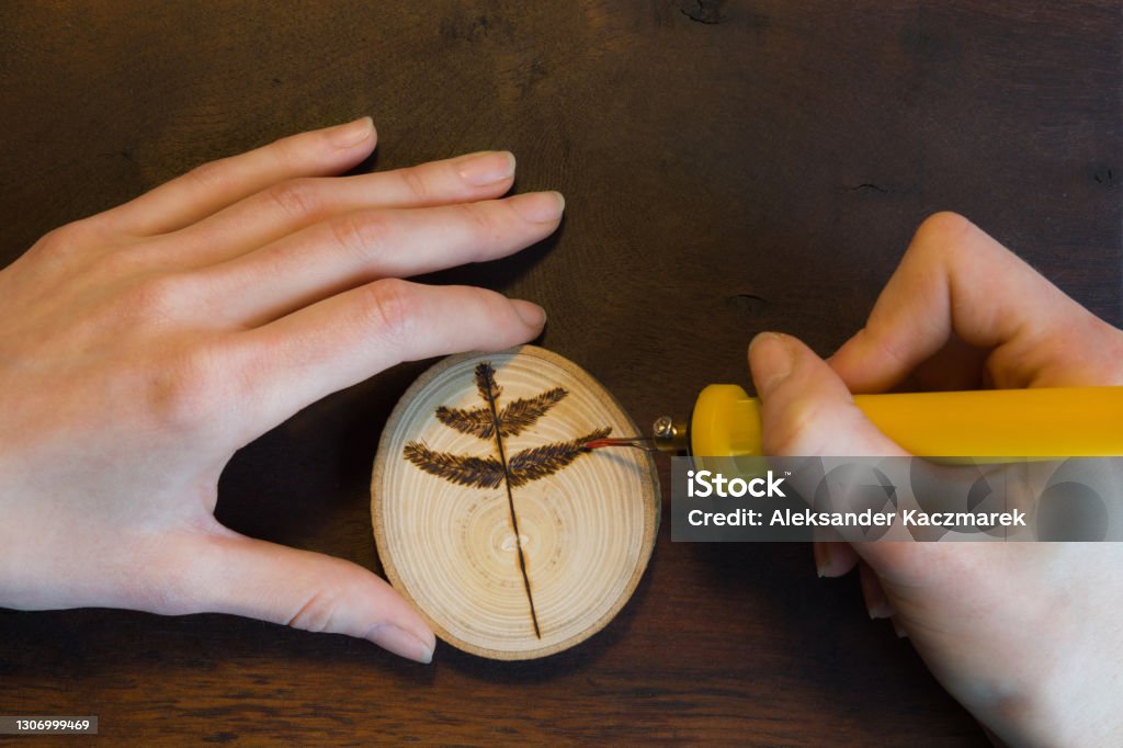 Close-up of female hands making a tree twig sign on wooden disc. Female hands burn out a tree twig sign with soldering iron on wooden disc. Pyrography Stock Photo