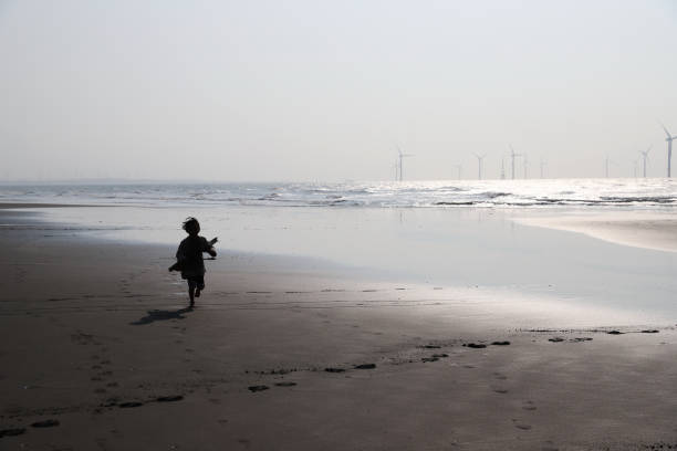 Young child wearing cap and shorts stands barefoot on Sandy Beach, looking thoughtfully at gentle ocean waves under bright daylight. Calm coastal scene captured from side angle