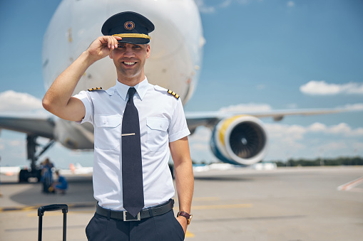 Cheerful young man airline worker touching captain hat and smiling while standing in airfield with airplane on background