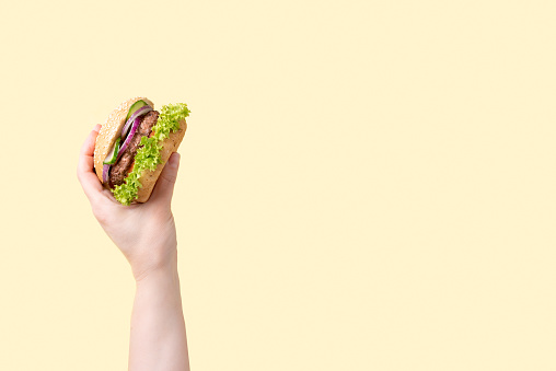 Female hand with tasty burger on yellow background, close up.