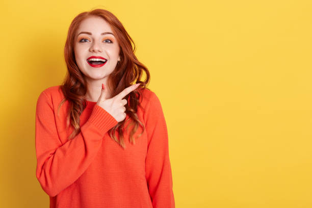 istock Attractive woman with happy expression pointing at copy space with index finger, dressed in casual orange sweater, models against yellow background, ginger female with excited expression. 1307020821