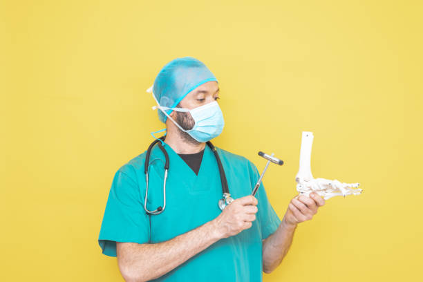 手術室の制服と聴診器を身に着けた外傷外科医は、すべての黄色の背景に足、右下肢の骨格モデルをチェックし��ています。 - senior adult human face male action ストックフォトと画像