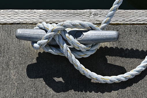 A white rope is tied to a cleat on a public pier made of wood and concrete. The day is sunny and we see hard shadows. Copy space.