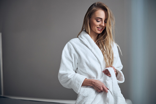 Smiling beautiful young woman with loose long hair tying the bathrobe belt at the waist