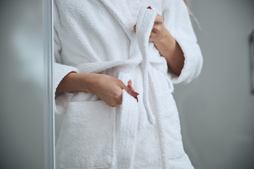 Cropped photo of a slim Caucasian woman tying the terry bathrobe belt at the waist in the bathroom