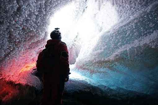 A guy in an ice cave with a lantern light. The caver descended into the ice cave. Snow stalactites and ice walls. In some places there are stones. Colored lantern beams. Bogdanovich Glacier, Almaty