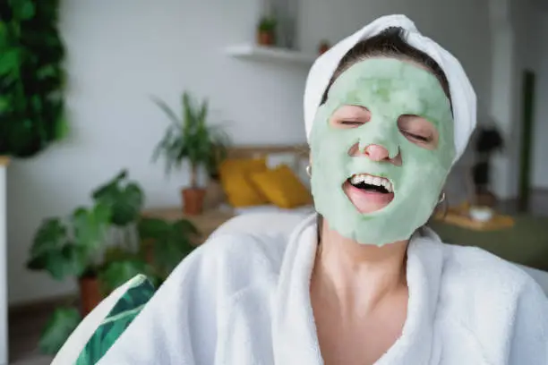 Photo of Beautiful young woman applies moisturizing green funny bubble face mask.Lying on couch. White bathrobe,towel.Self skin care,anti age morning treatment for wrinkles at home.Apartment with green plants