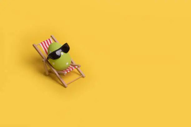Photo of Minimal  funny composition with Easter egg with sunglasses while sitting on deck chair on illuminating yellow background.