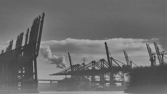 Container gantry cranes in the port of Hamburg in a misty industrial environment. A chimney in the background smokes and indicates great industie.