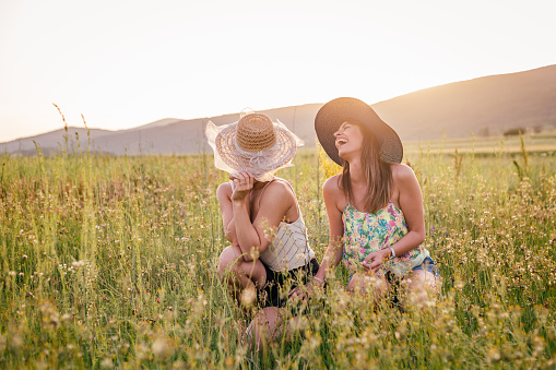 Two cheerful females sitting in the field with flowers on a warm day. Sun is setting behind them. They are holding hats.