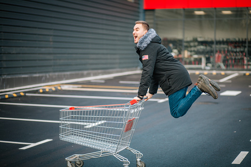 Smiling young man with a shopping cart jumping in front of a big shop