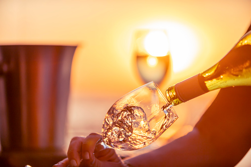 Close up of white wine being poured into a glass.  A second glass can be seen in the background. A woman is holding the glass and pouring the wine. Ice bucket and wine bottle with a romantic sunset.