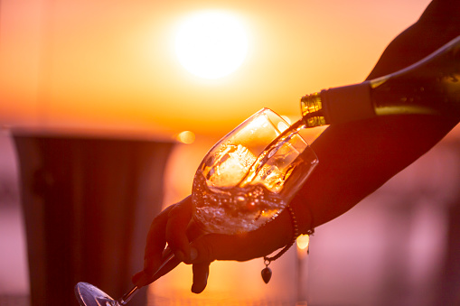 Close up of white wine being poured into a glass. A woman is holding the glass and pouring the wine. Ice bucket and wine bottle with a romantic sunset.