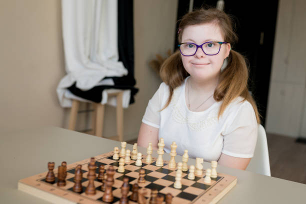 verticale de jeune fille de sourire avec le syndrome de down jouant aux échecs sur le plateau de jeu. gosses handicapés examinant l’échiquier à la maison. développement des capacités des enfants ayant des retards de développement - chess skill concentration intelligence photos et images de collection