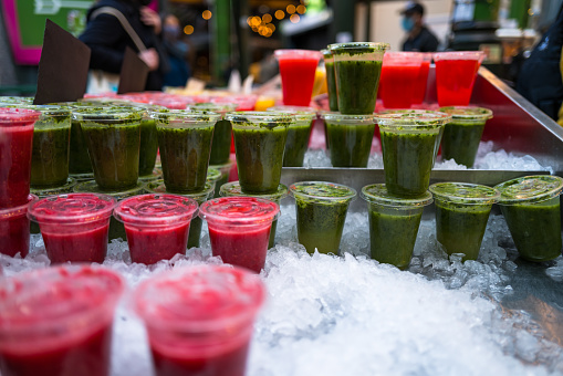 Close up color image depicting freshly made fruit juices and smoothies on display in a row and for sale at a food and drink market in London, UK. Selective focus on the plastic cups containing the fresh smoothies. In the background people are blurred out of focus. Room for copy space.