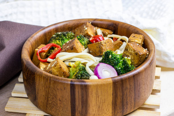 hausgemachter vegetarischer gebratener tofu mit udon-nudeln und gemüse (brokkoli, pfeffer und zwiebeln) in der holzschüssel auf leichtem küchenhintergrund - tofu chinese cuisine vegetarian food broccoli stock-fotos und bilder