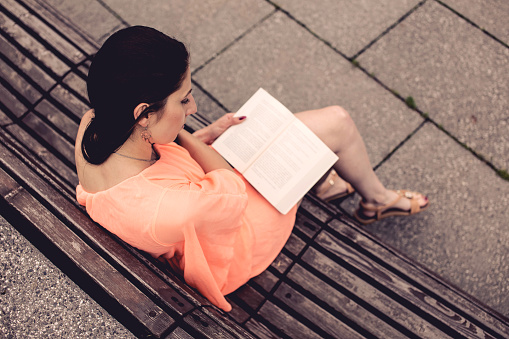 A girl with dark long hair wearing an orange dress is sitting on a park bench and reading a book