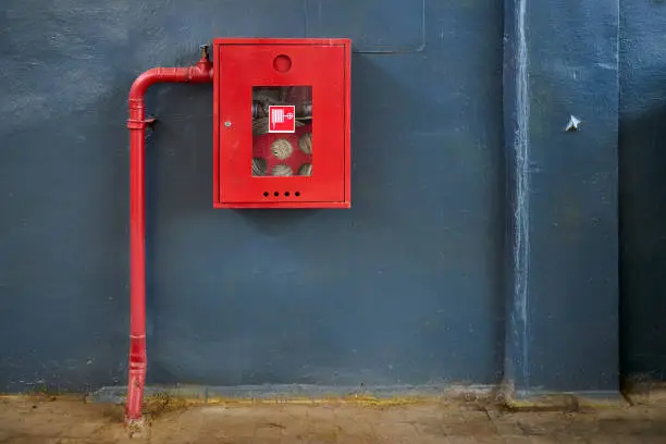 Fire hose in red box. Pipe roll for fire emergency in red metal cabinet on gray painted concrete wall with tiled floor as part of firefighting system of industrial production plant, with copyspace.